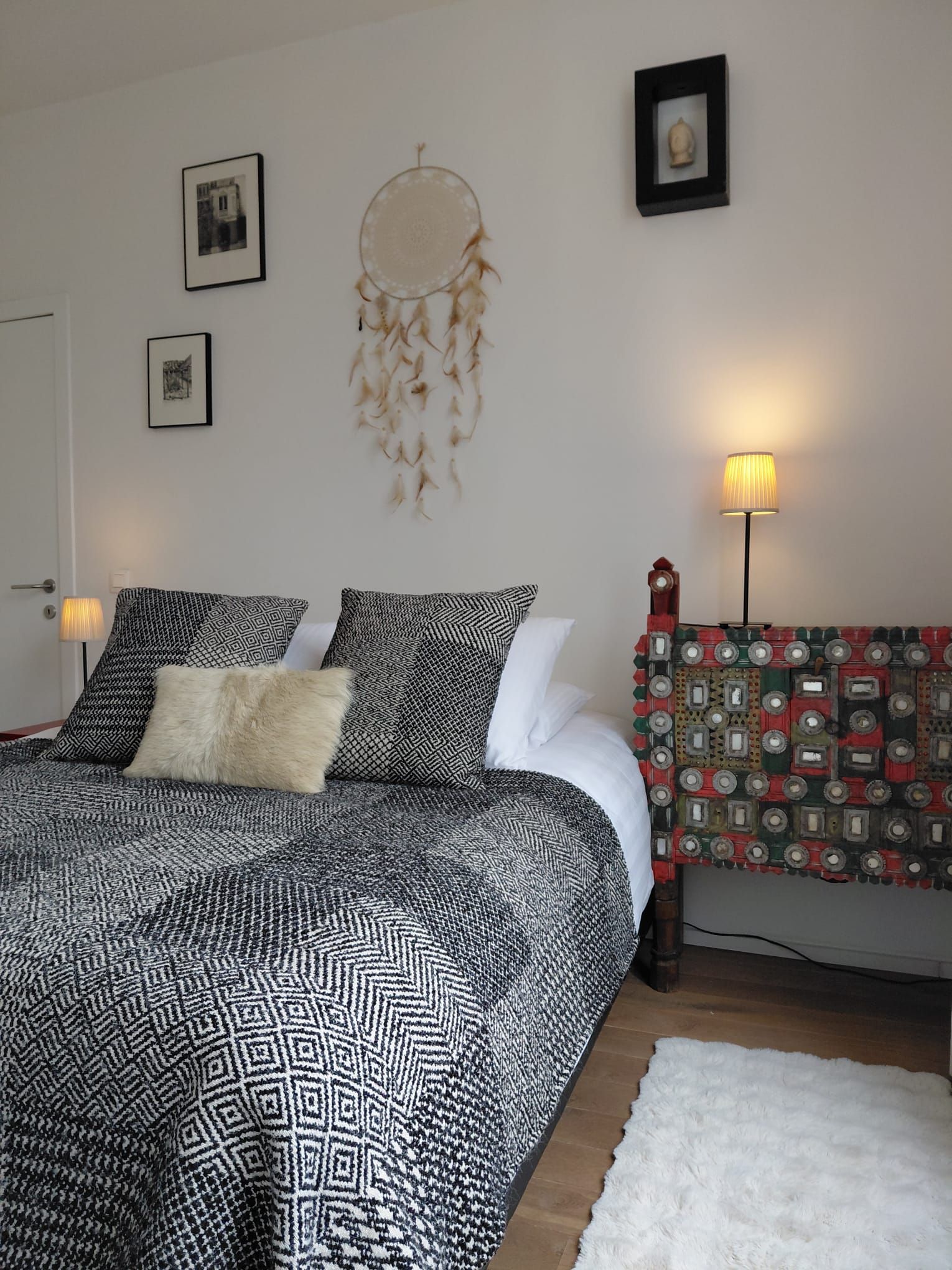 A bedroom with a patterned bedspread, pillows, a dreamcatcher, framed pictures, and a lamp.