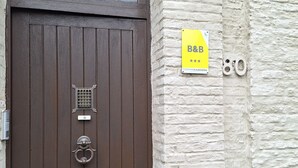 A brown wooden door with a window above it displaying a logo and the text "INGANG BUREEL". There is a yellow sign with "B&B" and three stars, and the number "80" on the wall next to the door.