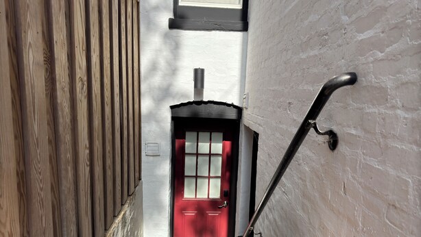 Un escalier étroit mène à une porte rouge, adossée à un mur blanc et bordée d’une clôture en bois d’un côté.