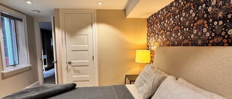 Un dormitorio con una cama grande, una mesita de noche, una lámpara de pared y una puerta que da a otra habitación.