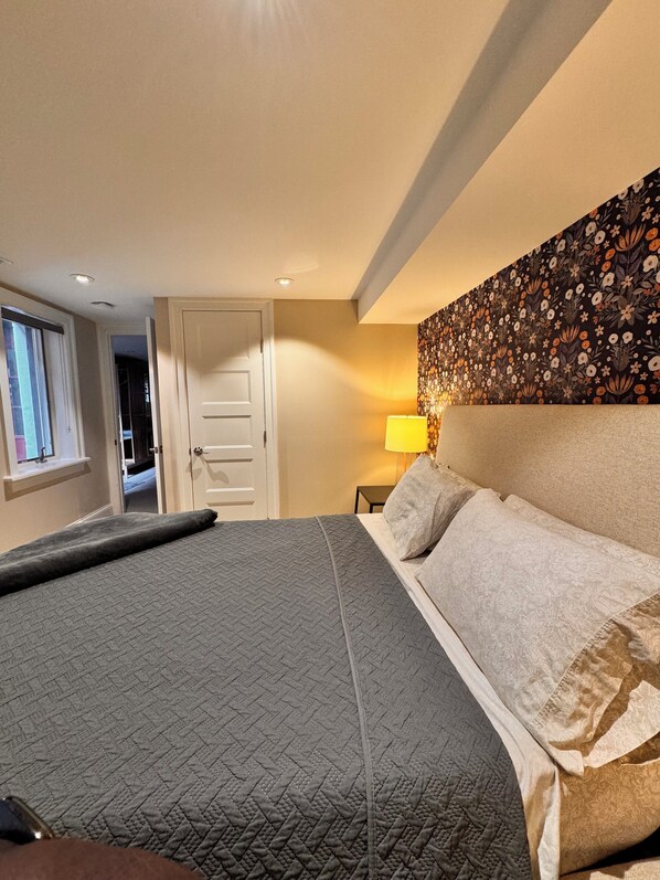 A bedroom with a large bed, a nightstand, a wall-mounted lamp, and a door leading to another room.