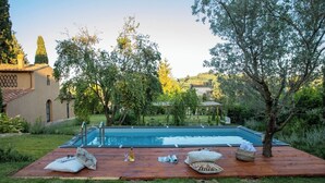 Piscina com deck de madeira, rodeada por vegetação e um edifício com telhado de azulejos.
