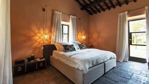 Quarto com uma cama grande, duas mesas de cabeceira, janela com cortinas e teto com vigas aparentes.