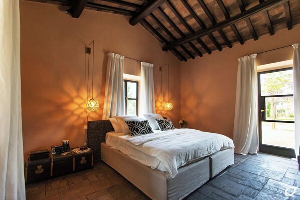 A bedroom with a large bed, two bedside tables, a window with curtains, and a ceiling with exposed beams.