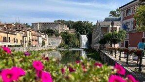 Ein Kanal mit einem Wasserfall, umgeben von historischen Gebäuden und blumenbepflanzten Promenaden.