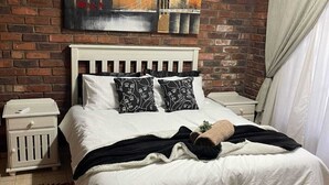 A bedroom with a brick wall, a white bed with patterned pillows, a red rug, and a ceiling fan.