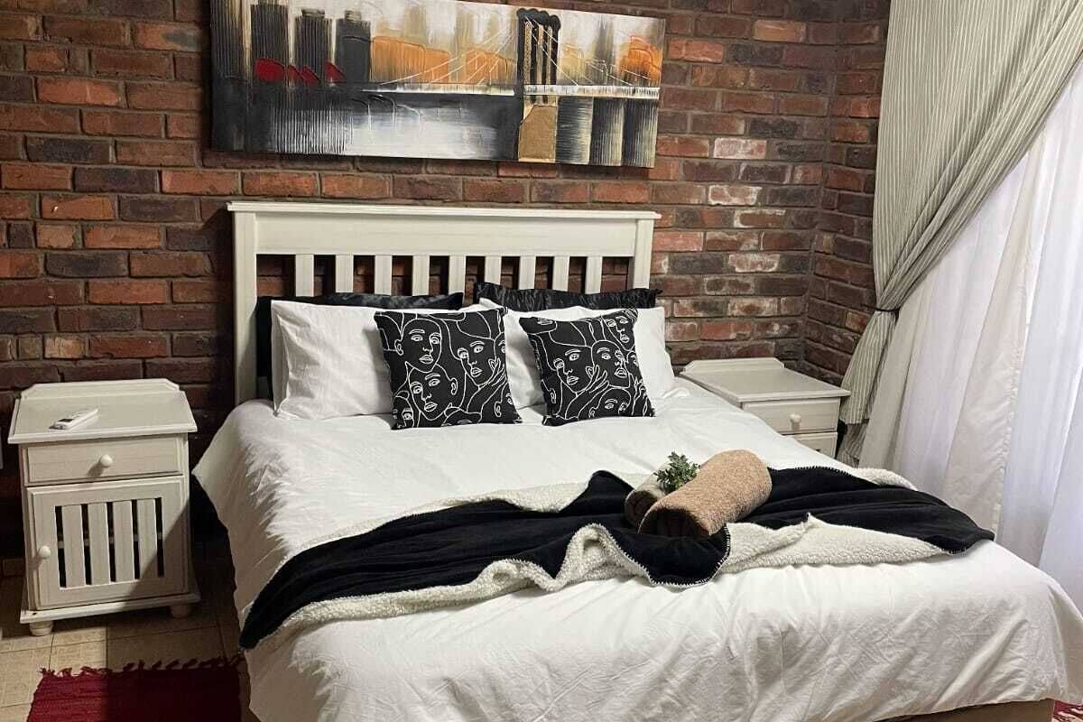 A bedroom with a brick wall, a white bed with patterned pillows, a red rug, and a ceiling fan.