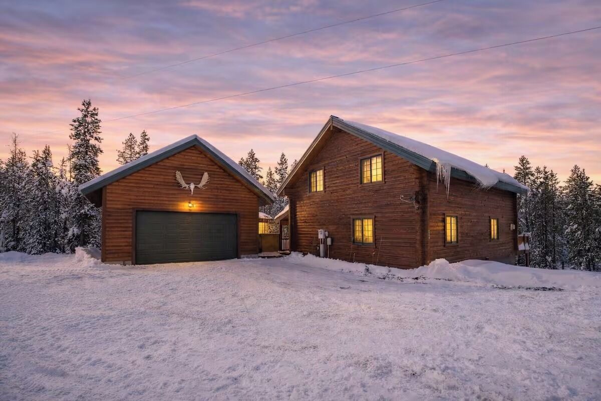 Kaksikerroksinen hirsihökkelö, jossa on autotalli ja lumipeitteinen katto, ja sen ympärillä on puita.