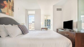 A hotel room with a large bed, a TV on a wooden dresser, and a city view through the window.