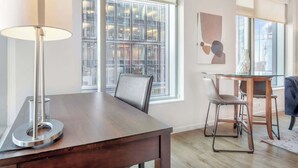 A modern dining area with a wooden table, metal chairs, and a glass-top table. Large windows provide natural light and views of the city.