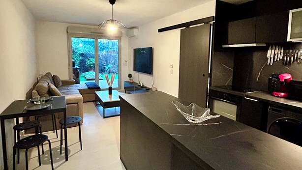 A modern kitchen with a central island, a dining area with a sofa and chairs, and a view of the outdoors through a large window.