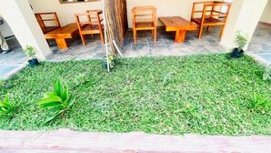 Un espace extérieur aménagé avec des bancs et des chaises en bois, un petit jardin avec de la pelouse et des plantes, et un sol carrelé.