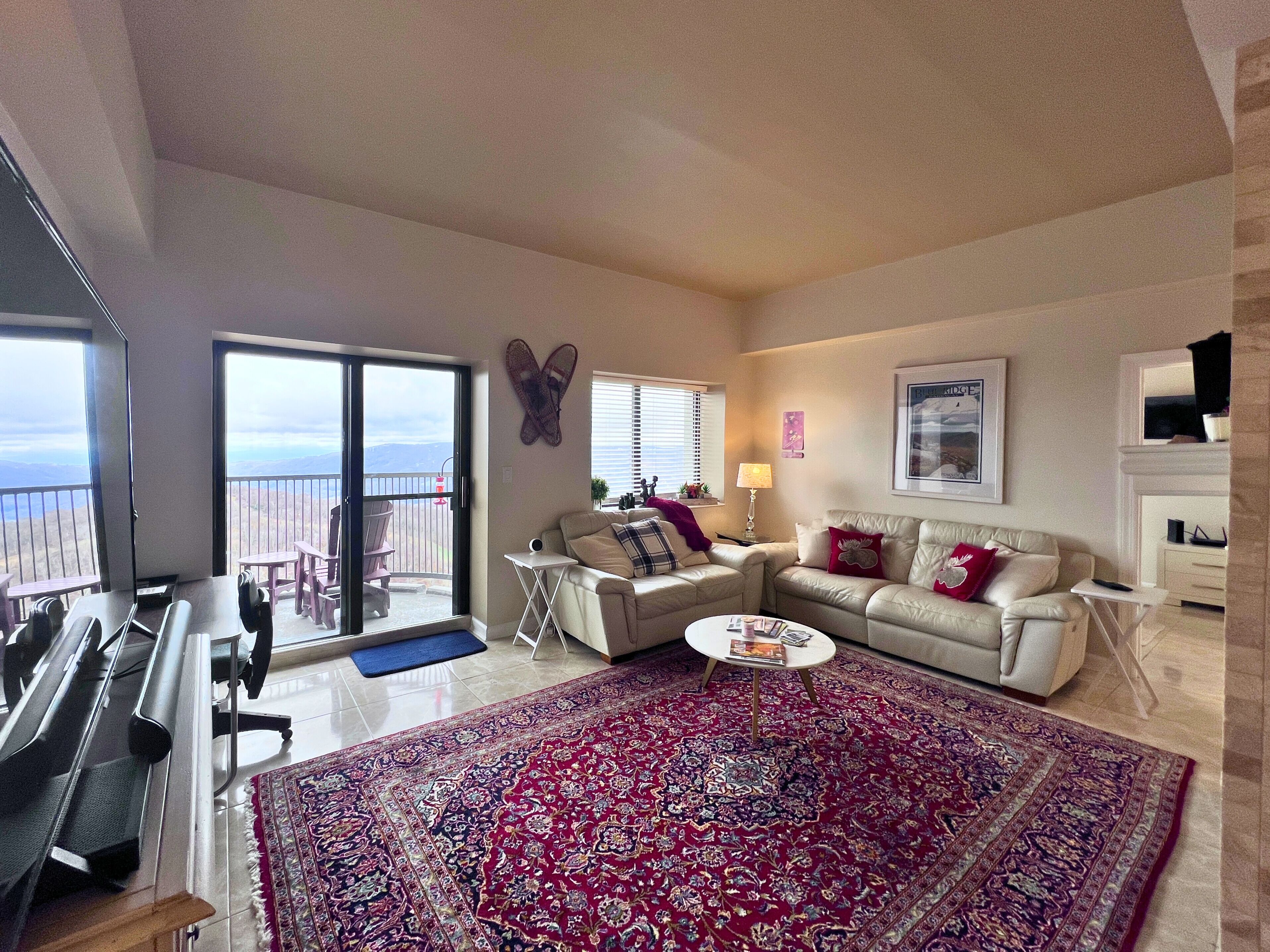 A spacious living room with a large red patterned rug, a white sofa with red and white pillows, a small round coffee table, and a balcony with a view of the mountains.
