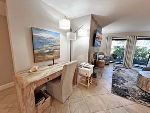 A modern living room with a wooden desk, a large painting, a flat-screen TV, and a comfortable seating area with a view to the outdoors.