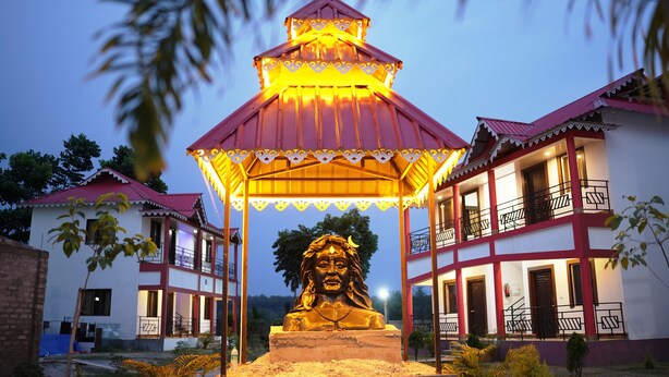 Un bâtiment avec un toit rouge et des murs blancs, une statue dorée devant, et un kiosque illuminé de lumières jaunes.