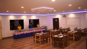 Un espace repas avec des tables et des chaises en bois, une table de buffet avec des récipients contenant de la nourriture et des luminaires au plafond.