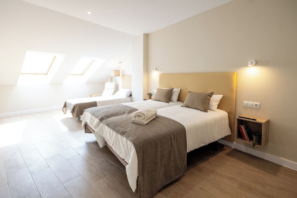 Una habitación de hotel moderna con dos camas, una mesita de noche con lámpara, un estante pequeño con libros y tragaluces.