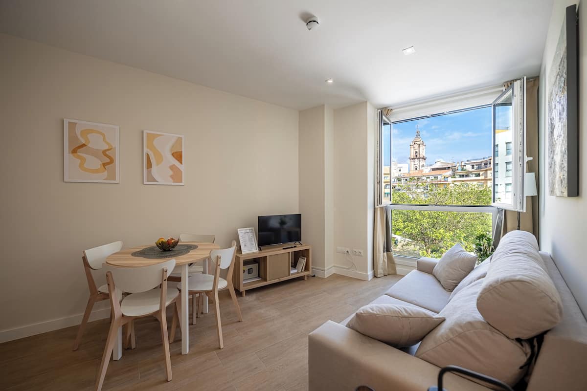 A modern living room with a dining area, a flat-screen TV, and a large window offering a city view.