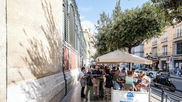 Une scène de rue avec des tables et des chaises installées à l’extérieur, sous un auvent, une enseigne indiquant « mercados de Málaga », et des bâtiments en arrière-plan.