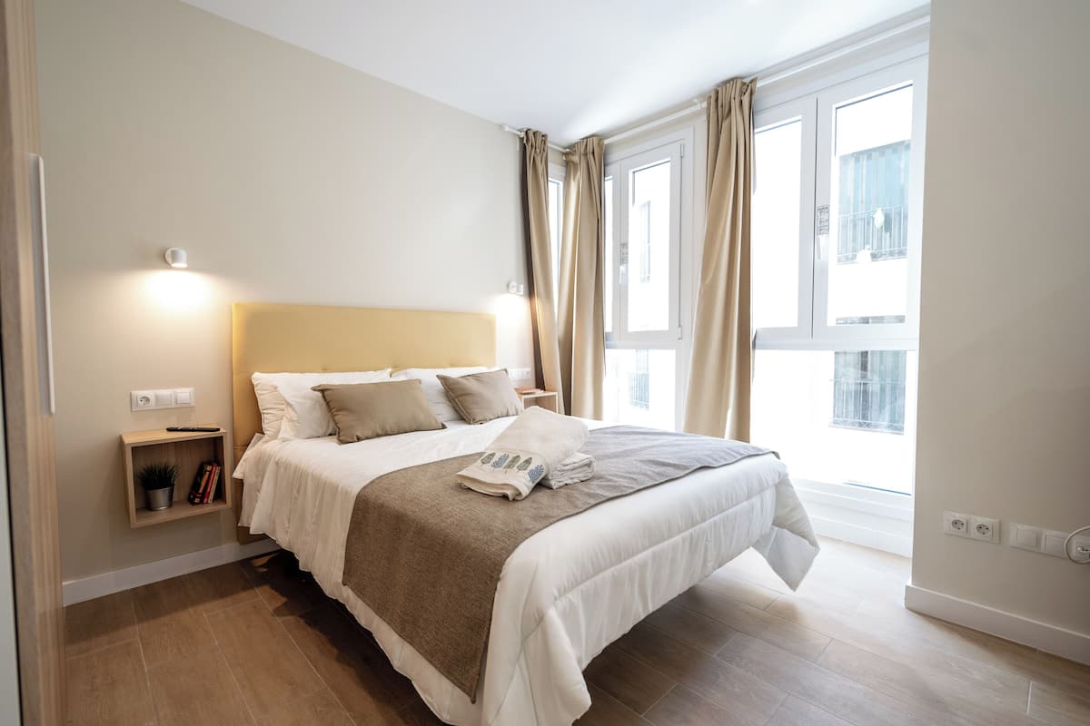 A neatly made bed with a headboard, a bedside table with a lamp, and a window with curtains.
