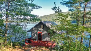 A cabin with a red door and a porch, surrounded by trees and a lake. - Charming boathouse for two with deck overlooking Lake Kanasatka (Moultonborough)