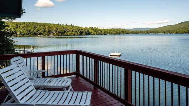 A deck with white lounge chairs overlooking a calm lake, surrounded by lush greenery and distant hills. - Charming boathouse for two with deck overlooking Lake Kanasatka (Moultonborough)
