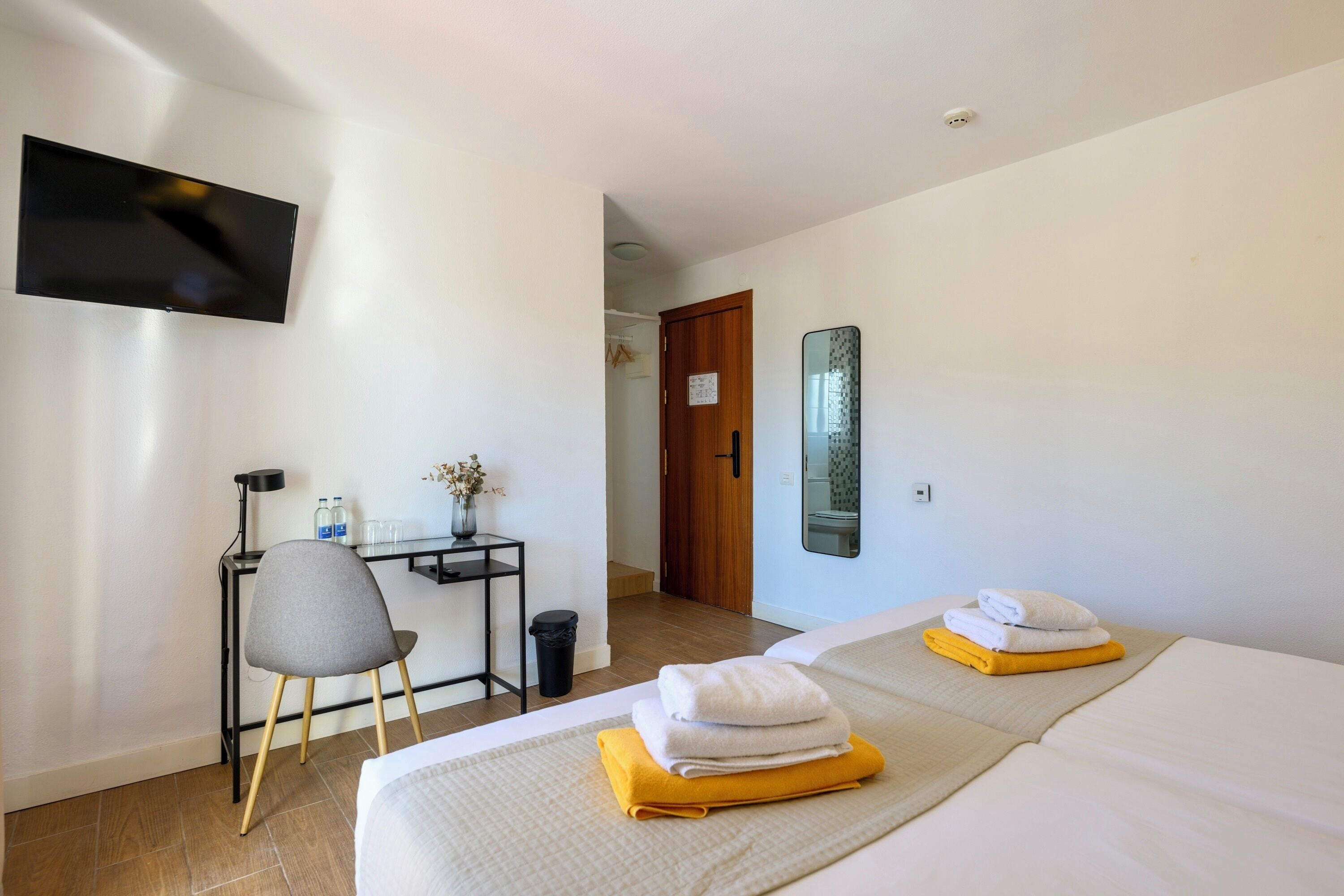 A hotel room with two beds, a desk with a chair, a TV mounted on the wall, and a mirror.