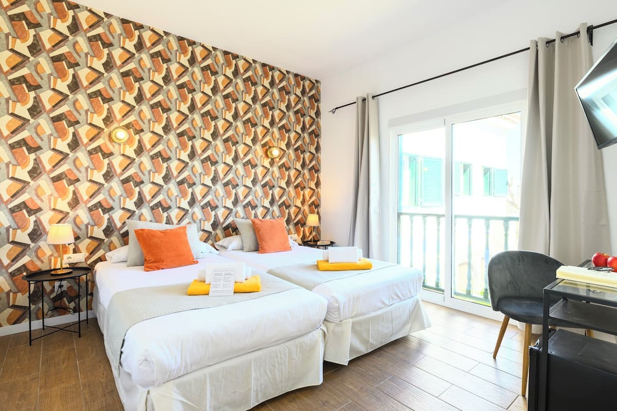 Een hotelkamer met twee bedden, een behang met patroon, een balkon met uitzicht en een bureau met stoel.
