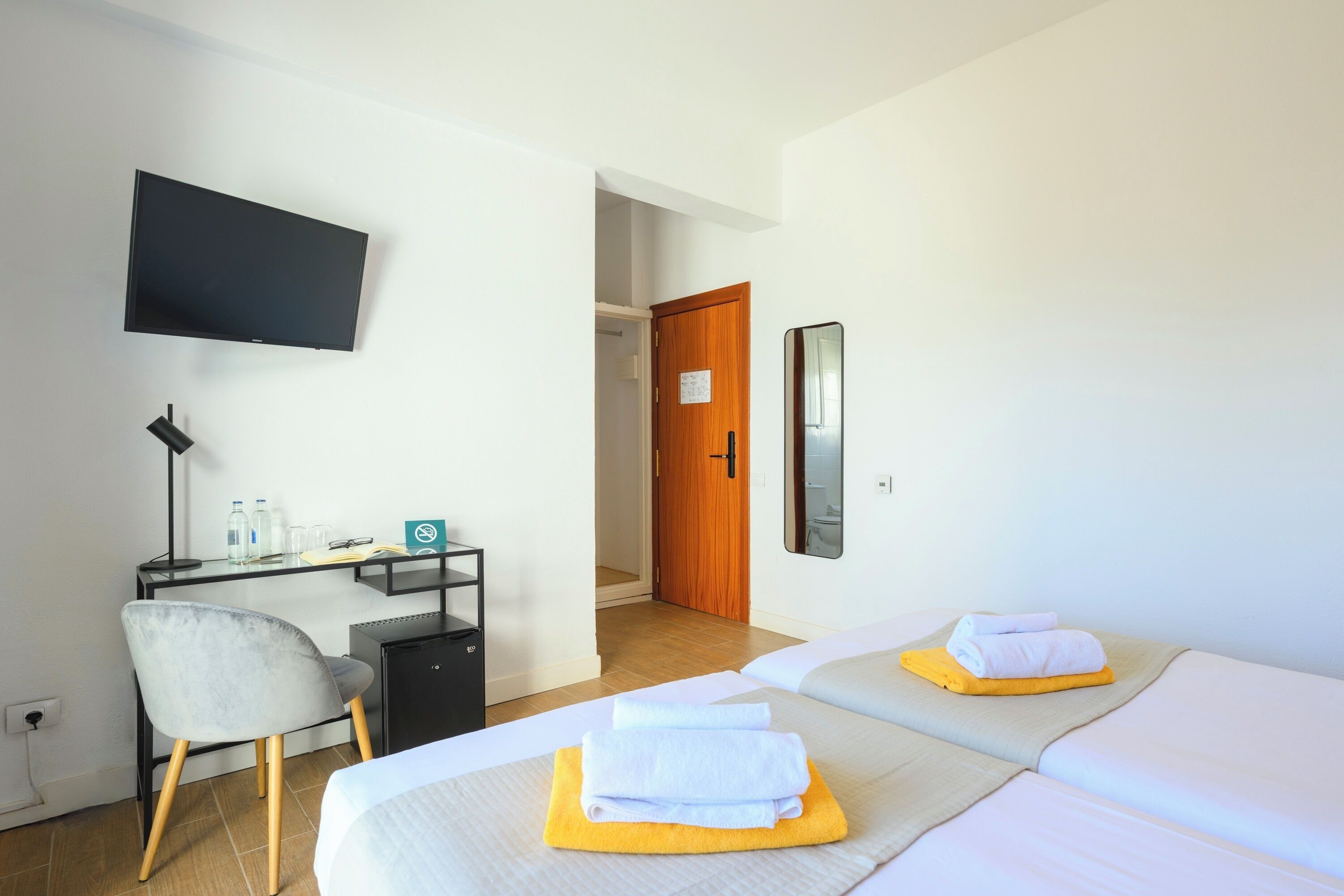 A hotel room with two beds, a desk, a chair, a TV, and a door leading to another room.