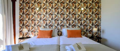 Quarto de hotel com duas camas, cada uma com roupa de cama branca e almofadas laranja. Há duas mesas de cabeceira com luminárias. As paredes têm um papel de parede com padrão geométrico.