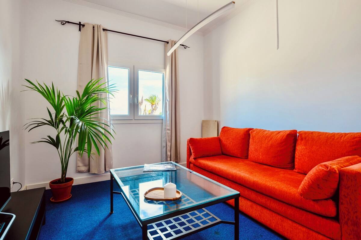 Een kamer met een glazen salontafel, een rode bank, een potplant en een raam met gordijnen.