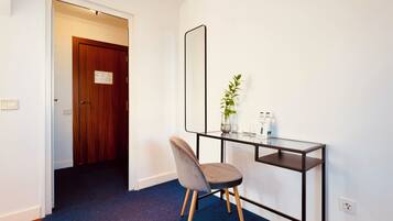 A room with a dark blue carpet, a wooden door, a glass desk with a mirror, a grey chair, a small plant, and a few toiletries on the desk.