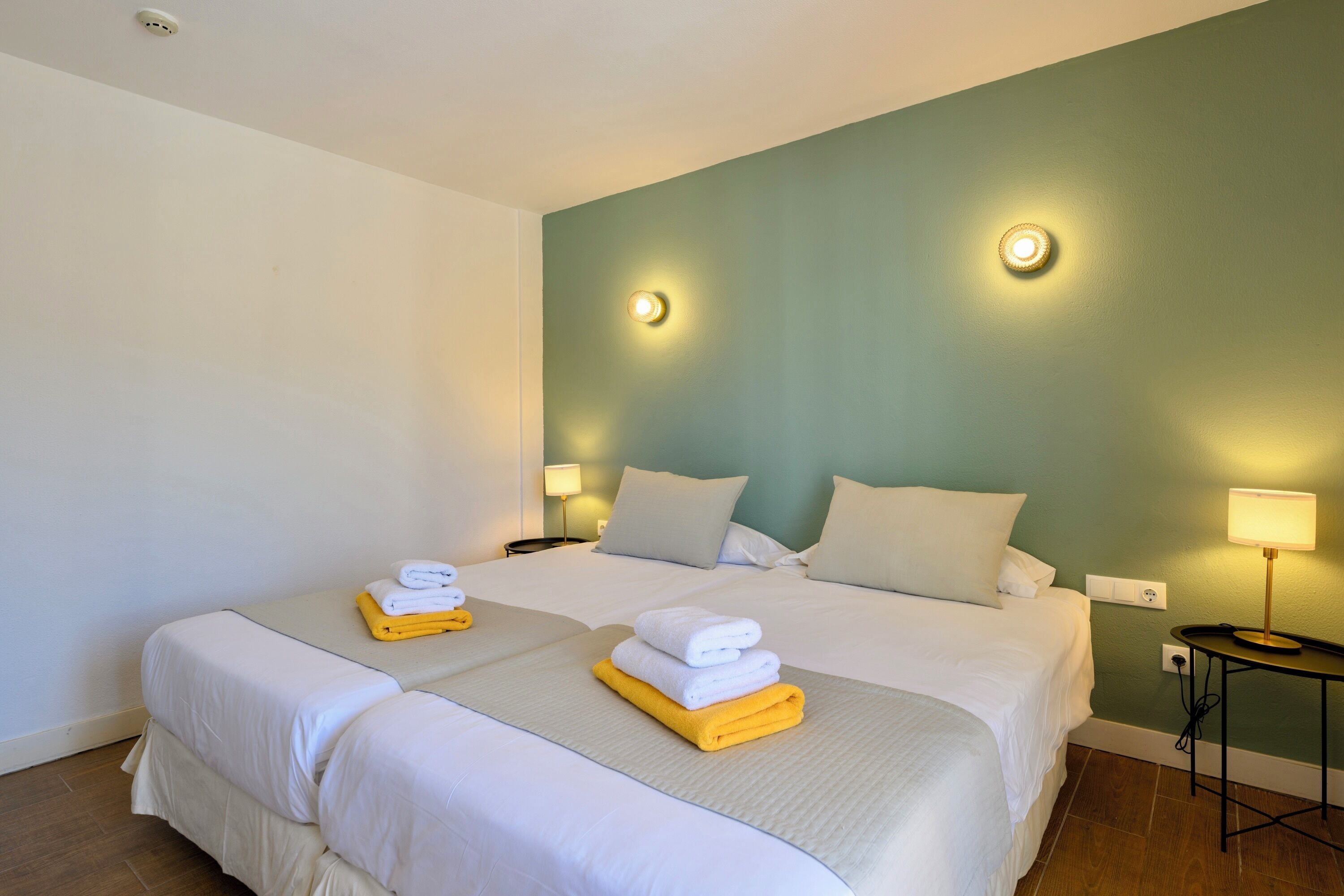 A hotel room with two beds, each with white linens and a yellow towel. There are two wall-mounted lamps and a small table with a lamp.