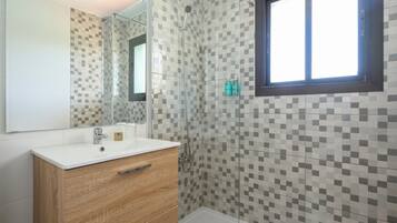 Een badkamer met een betegelde douchewand, een witte wastafel en een houten wastafelmeubel.