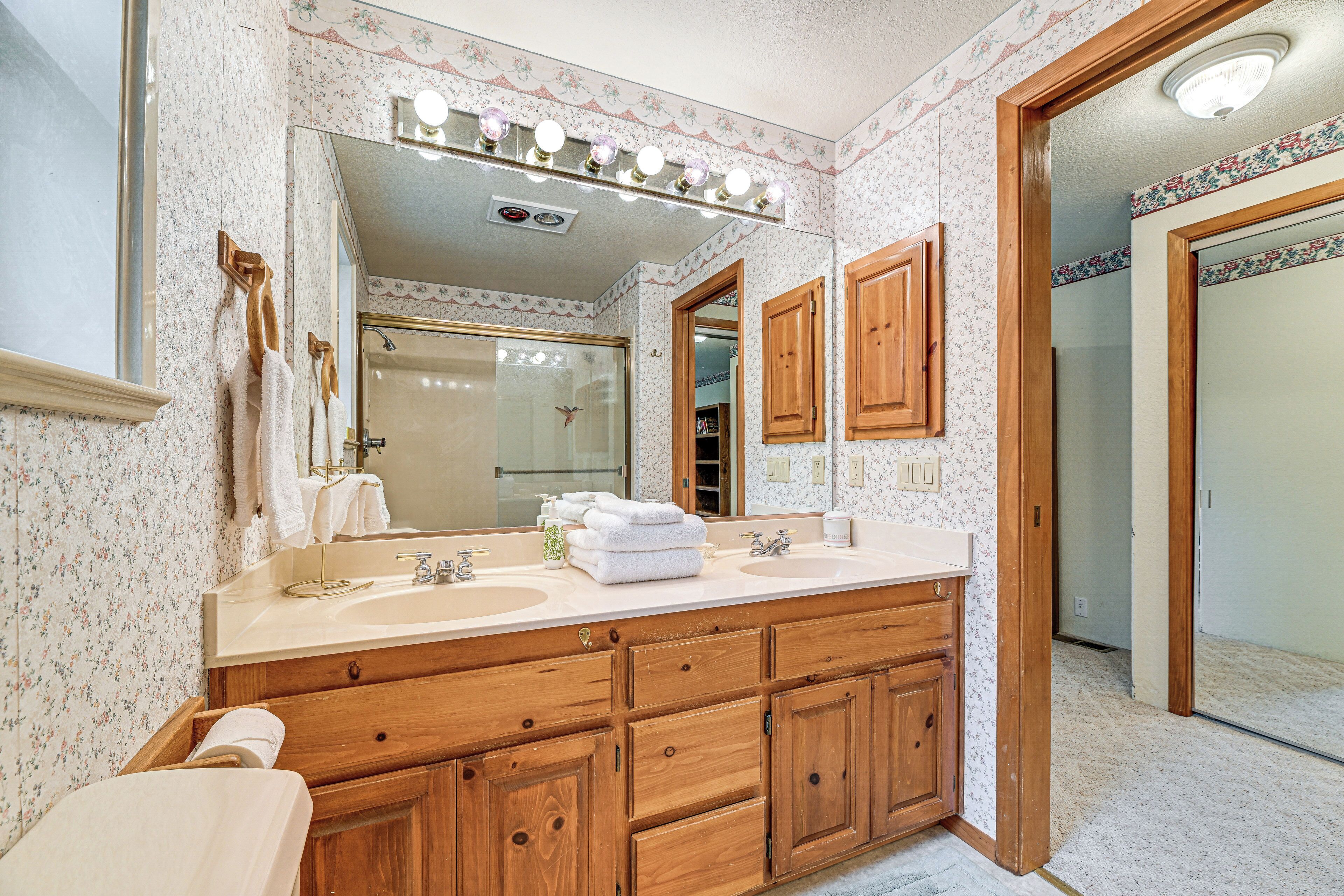 Une salle de bain avec un meuble-lavabo en bois, deux vasques, un grand miroir avec éclairage intégré et une douche visible au fond.