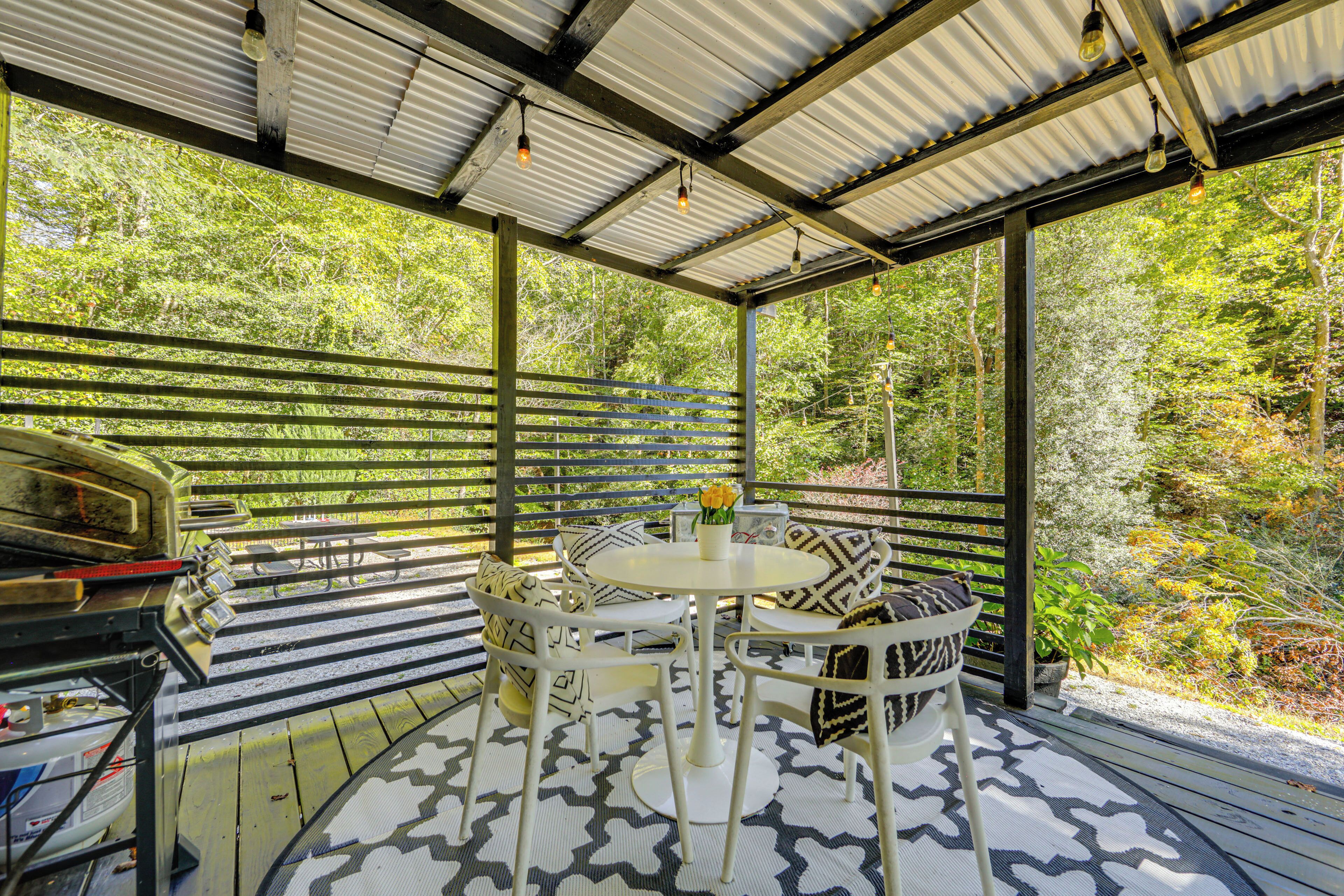 A covered outdoor seating area with a round table and chairs, surrounded by a metal railing, overlooking a forested area.