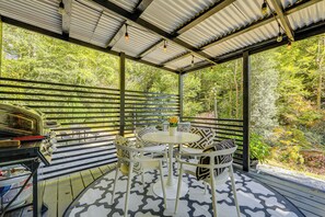 A covered outdoor seating area with a round table and chairs, surrounded by a metal railing, overlooking a forested area. - Blue Ridge Mountain Tiny Home: Escape to Nature! (Old Fort)