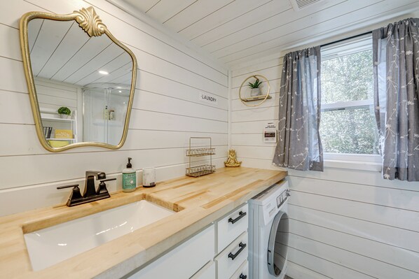 A bathroom with a wooden vanity, a sink, a mirror, a towel rack, and a window with curtains. - Blue Ridge Mountain Tiny Home: Escape to Nature! (Old Fort)