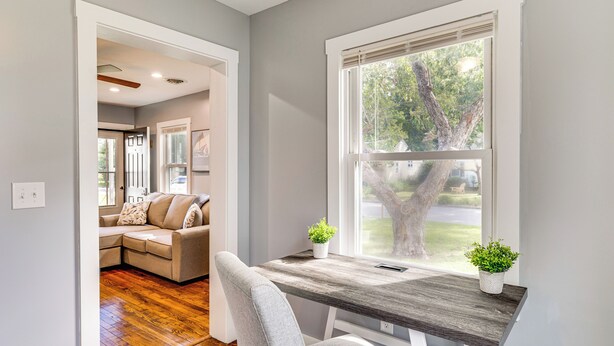 A modern interior with a wooden dining table, a grey sofa, and a window with a view of a tree and street. - 3 Mi to Camp Lejeune: Home w/ Fire Pit & Grill! (Jacksonville)