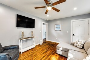 A living room with a ceiling fan, a flat-screen TV, a wooden floor, a grey sofa, and a white console table. - 3 Mi to Camp Lejeune: Home w/ Fire Pit & Grill! (Jacksonville)