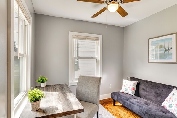 Une pièce avec une table en bois, un fauteuil gris, un canapé gris agrémenté de coussins à motifs floraux, un ventilateur de plafond et une fenêtre avec des stores.