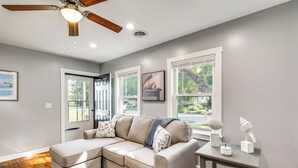 A modern living room with a sectional sofa, a ceiling fan, and large windows offering a view of the outdoors. - 3 Mi to Camp Lejeune: Home w/ Fire Pit & Grill! (Jacksonville)