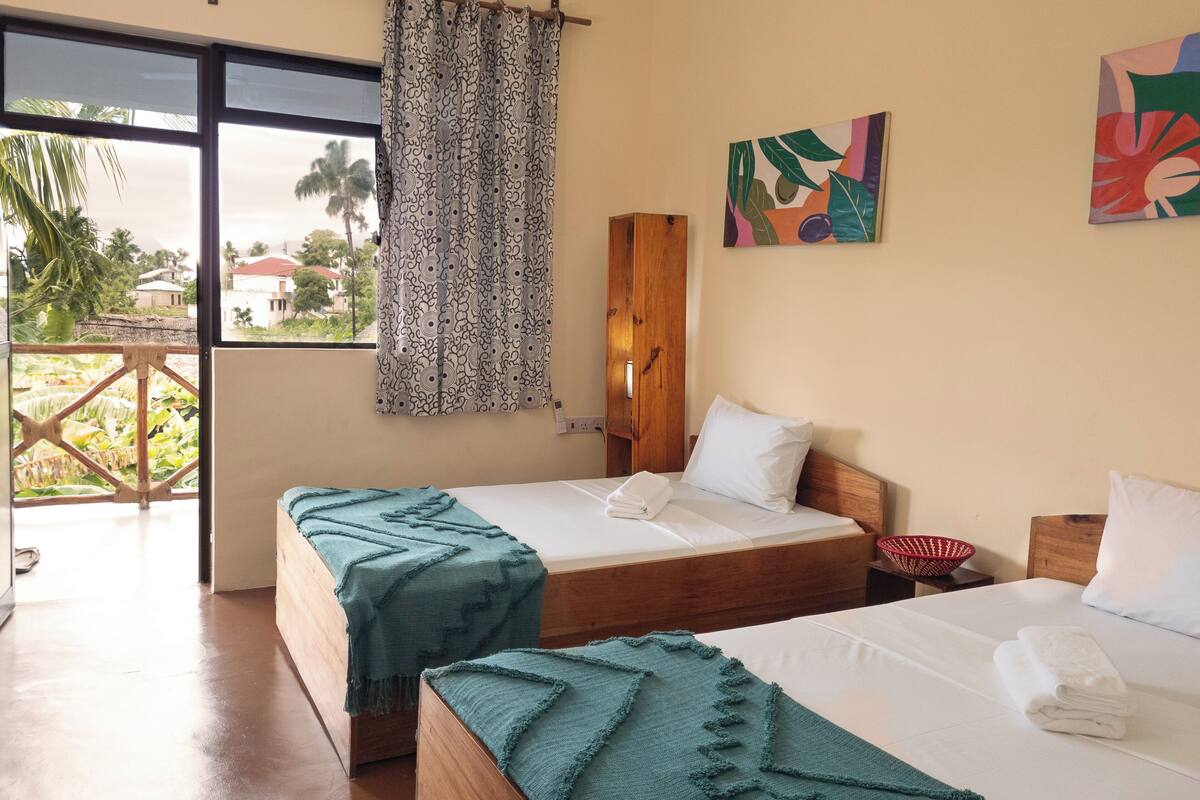 A room with two beds, a window with a view of palm trees, a wooden headboard, and a colorful abstract painting.