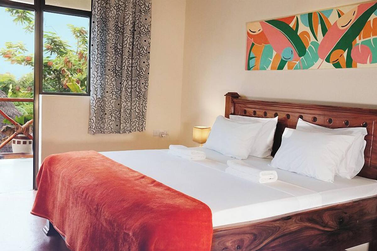 A hotel room with a large bed, a wooden headboard, a window with curtains, and a tropical view.