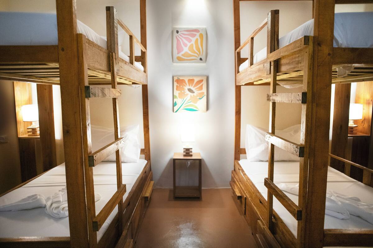 A corridor with bunk beds on either side, a small table with a lamp, and two framed floral artworks on the wall.