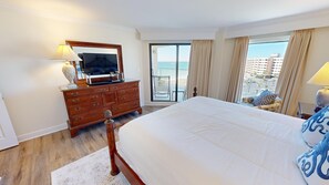 Habitació d'hotel amb un llit gran, una petarrera amb un televisor, un balcó amb vista al mar i un paisatge urbà al fons.