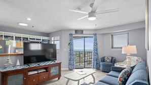 A modern living room with a flat-screen TV, a wooden entertainment unit, a blue sofa, a small table, and a balcony with a view.