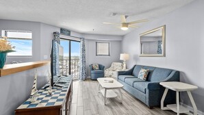 A room with a checkered floor, a blue sofa, a white table, a ceiling fan, and a view of the ocean.