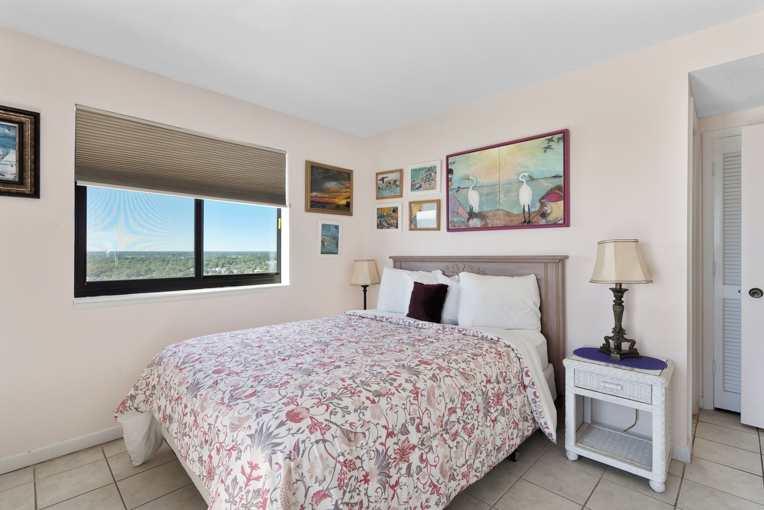 A bedroom with a large bed, a window with blinds, a nightstand with a lamp, and artwork on the walls.