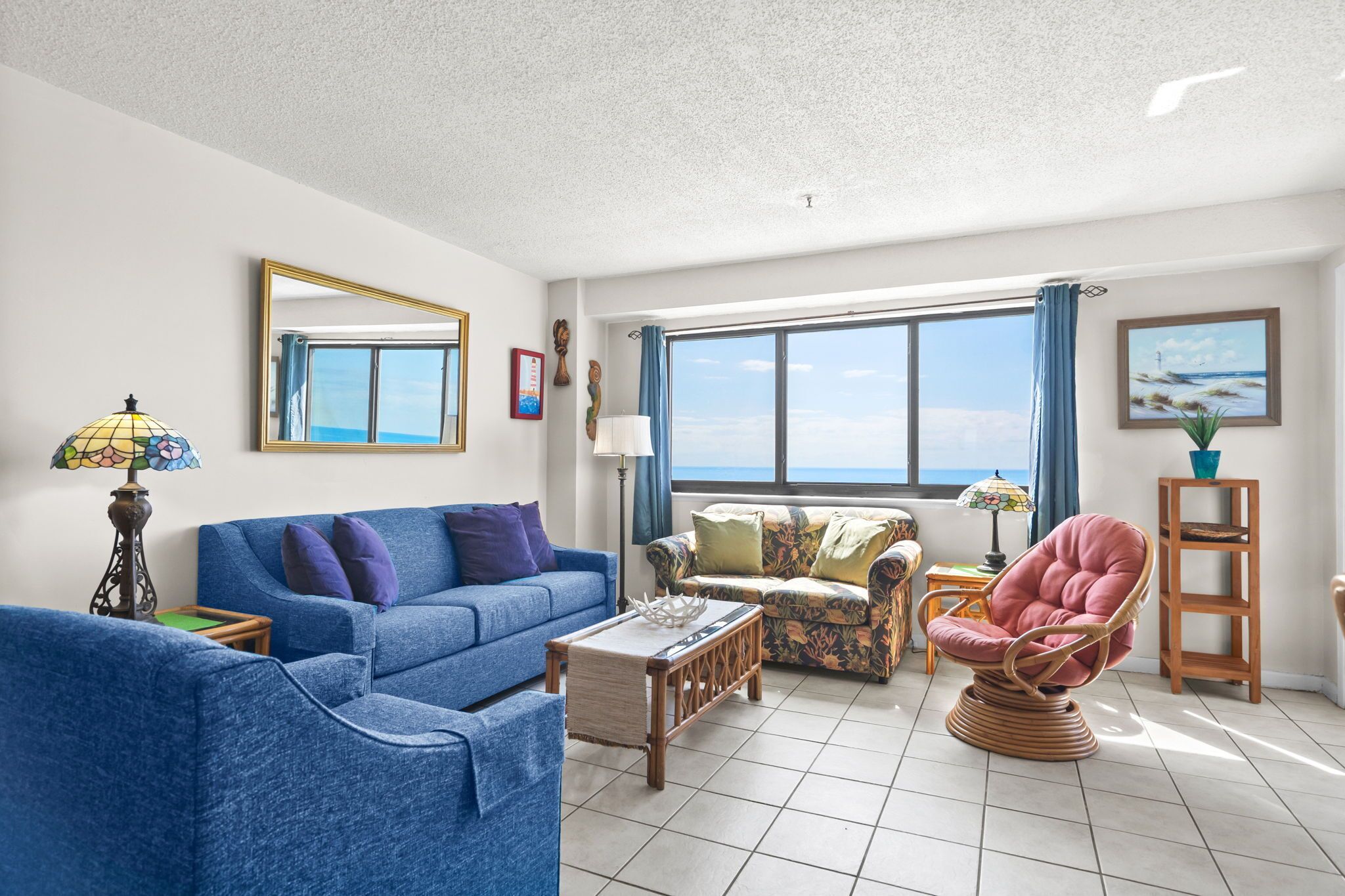 A living room with a blue sofa, a patterned sofa, a wicker chair, a small table, and a large window with a view.